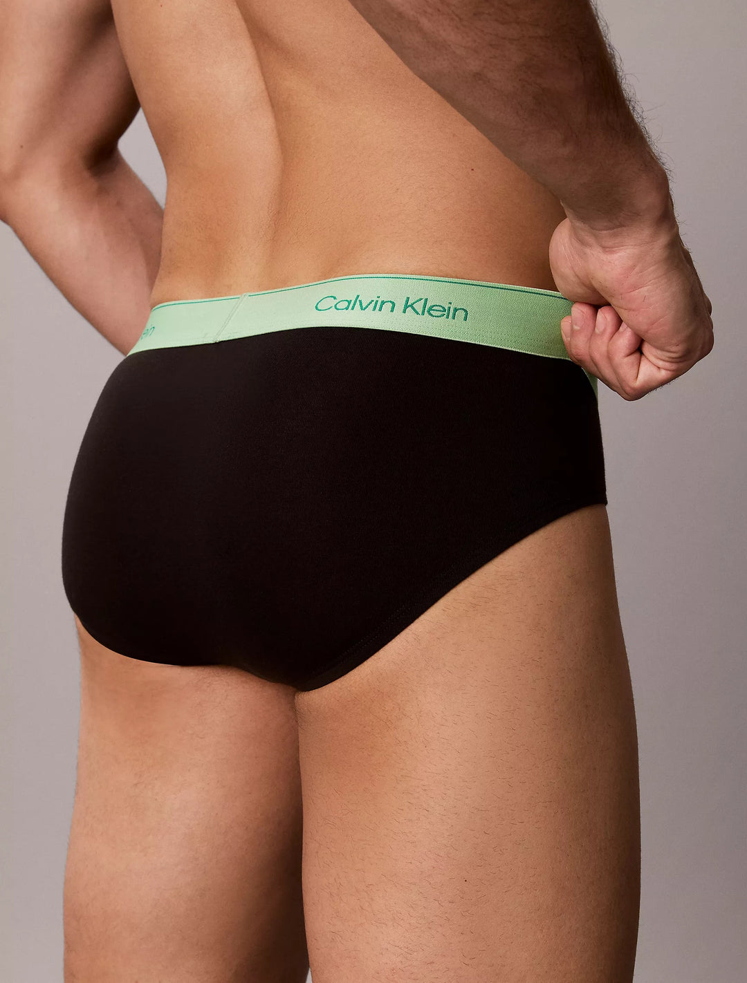 Close-up back view of Calvin Klein Pride brief showing mint green logo waistband on black, grey studio background